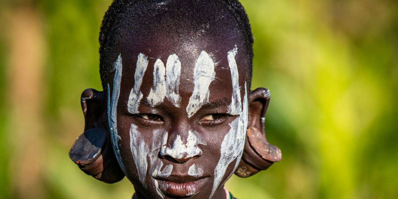 Kind in Äthiopien mit weißer Farbe im Gesicht und großen Ohrpflöcken in den Ohrläppchen