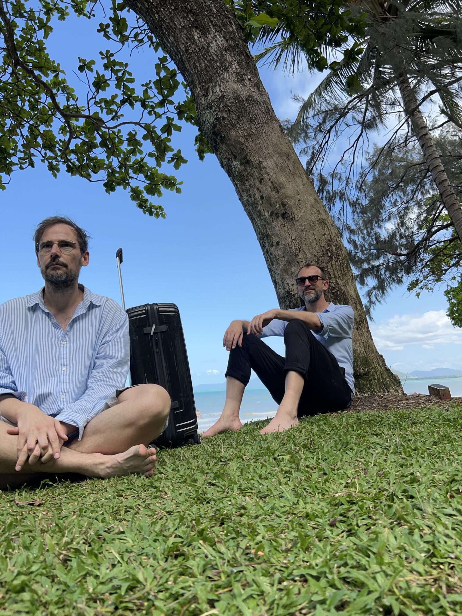 Jochen und Michael sitzen vor einem Strand mit Koffer in Port Douglas, Queensland