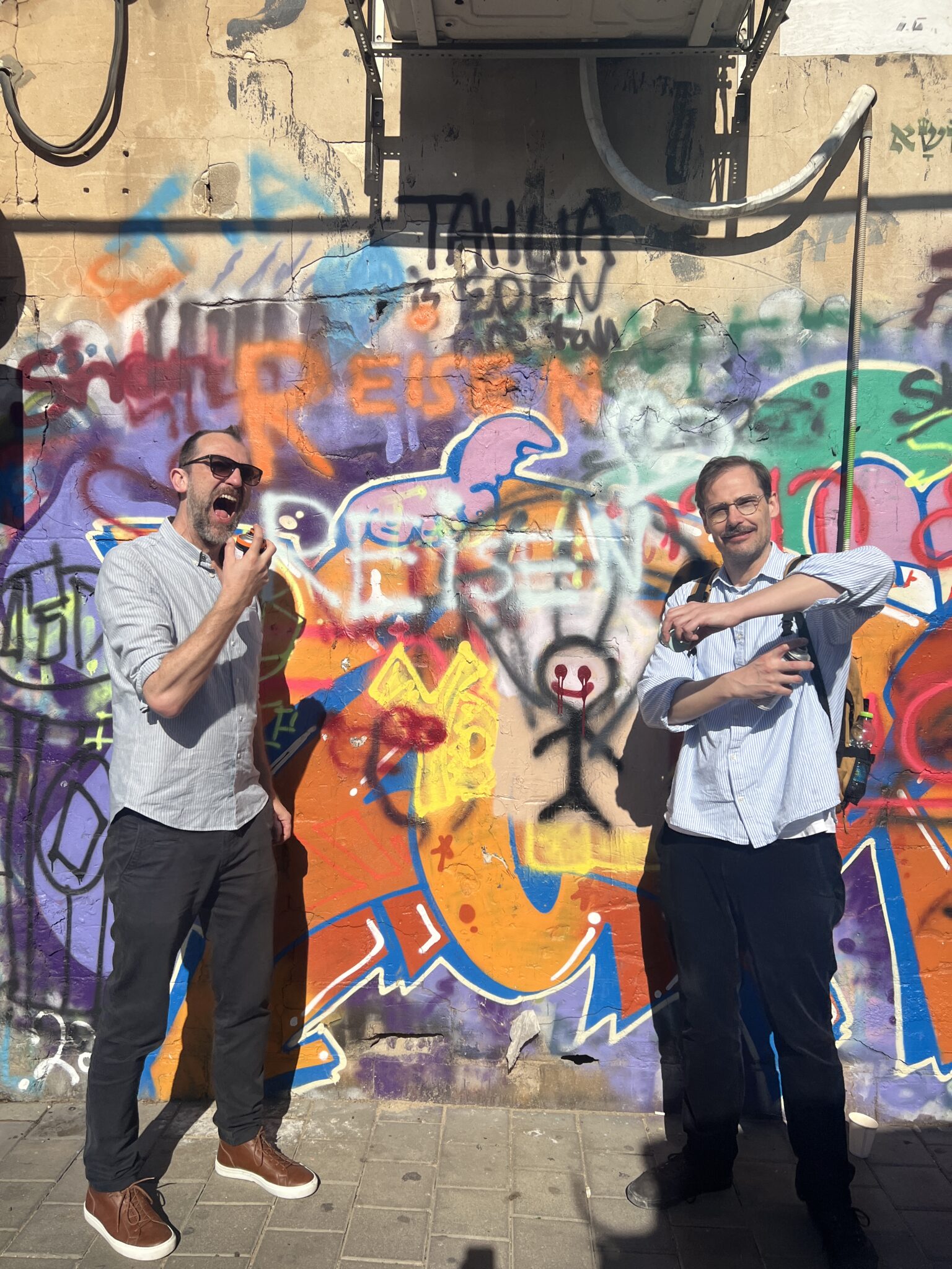 Michael und Jochen haben Reisen Reisen auf eine Graffiti-Wand gesprüht und posen witzig davor. Michael tut so als würde er sich mit der Dose in den Mund sprühen, Jochen benutzt sie scheinbar als Deospray