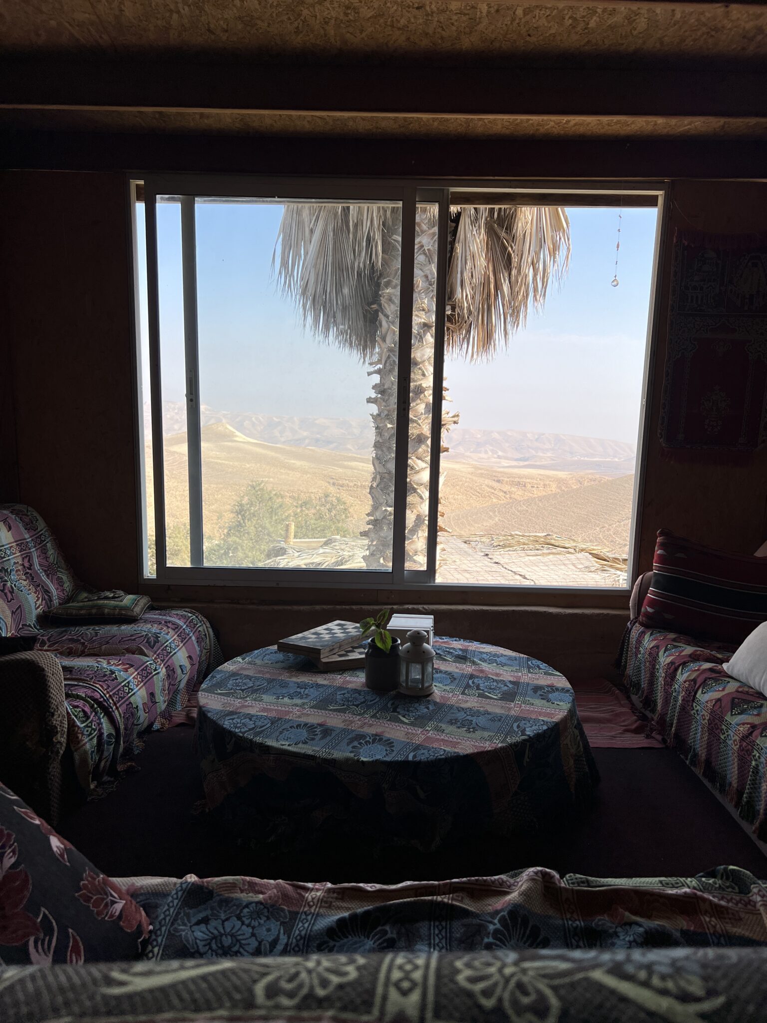 Die Bleibe eines Aussteigers, der in Arad Retreats anbietet. Ein gemütlicher Raum mit sofas und rundem Tisch in der Mitte, ein großes Fenster mit Palme davor und Blick auf die Wüste